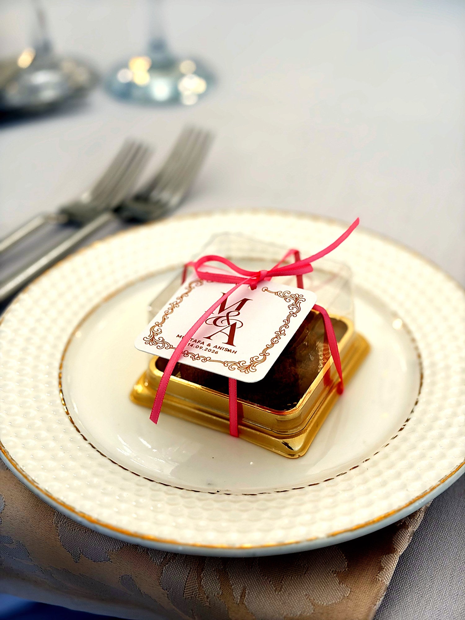 An Enchanted wedding favour: a gold box tied with pink ribbon and a personalised tag, on a guest's plate